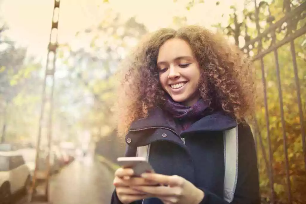 NBAD Balance Inquiry UAE – 7 No-Fee Methods in 2025 23 Young woman with curly hair smiles while texting on her smartphone outdoors. Autumn setting.