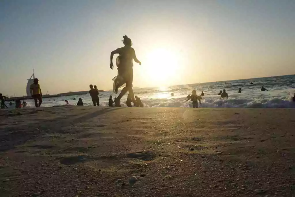 People enjoying water activities during sunset at Jumeirah Beach, Dubai with Burj Al Arab in background.