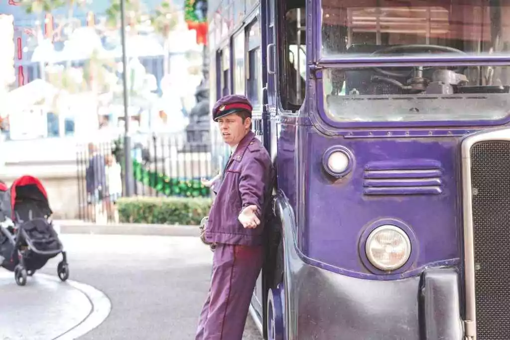 Conductor in uniform posing beside a vintage purple bus on a city street.