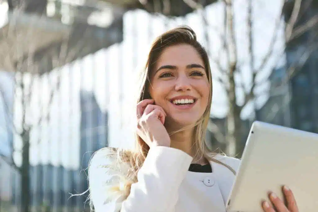 سيدة أعمال واثقة من نفسها تستخدم جهازها اللوحي والهاتف، وتبتسم في الهواء الطلق تحت ضوء الشمس.