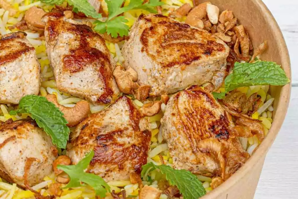 Closeup of grilled chicken pieces on seasoned rice with cashews and mint leaves in a bowl.