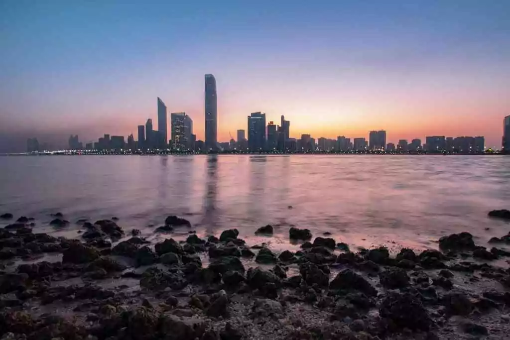 Stunning Abu Dhabi cityscape at sunrise with calm ocean foreground and vibrant sky.