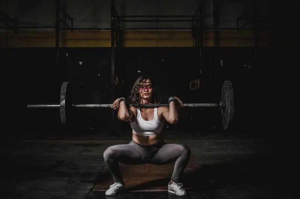 A strong female weightlifter squats with a barbell in a dimly lit gym, showcasing power and fitness.