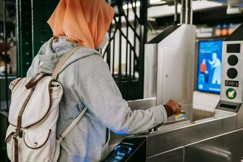 منظر خلفي لامرأة مسلمة ترتدي زيًا غير رسمي وحجابًا مع حقيبة ظهر تستخدم بطاقة السفر لعبور بوابة المترو في النهار