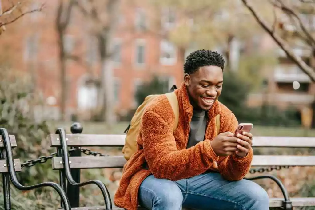 A cheerful young man in warm clothes smiling while using his phone on a bench in a city park.