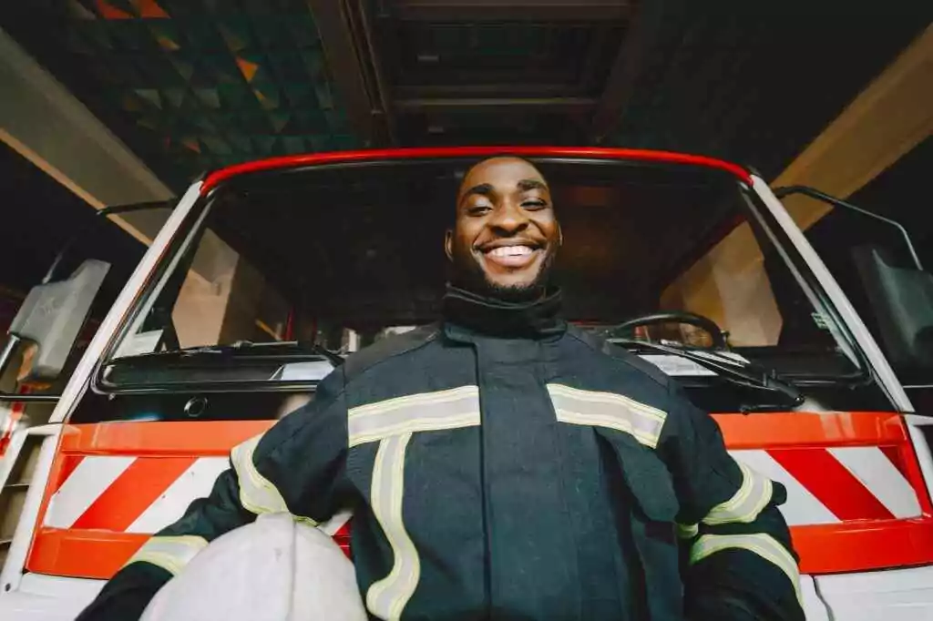 Smiling firefighter in safety gear standing in front of a fire truck, conveying bravery and readiness.