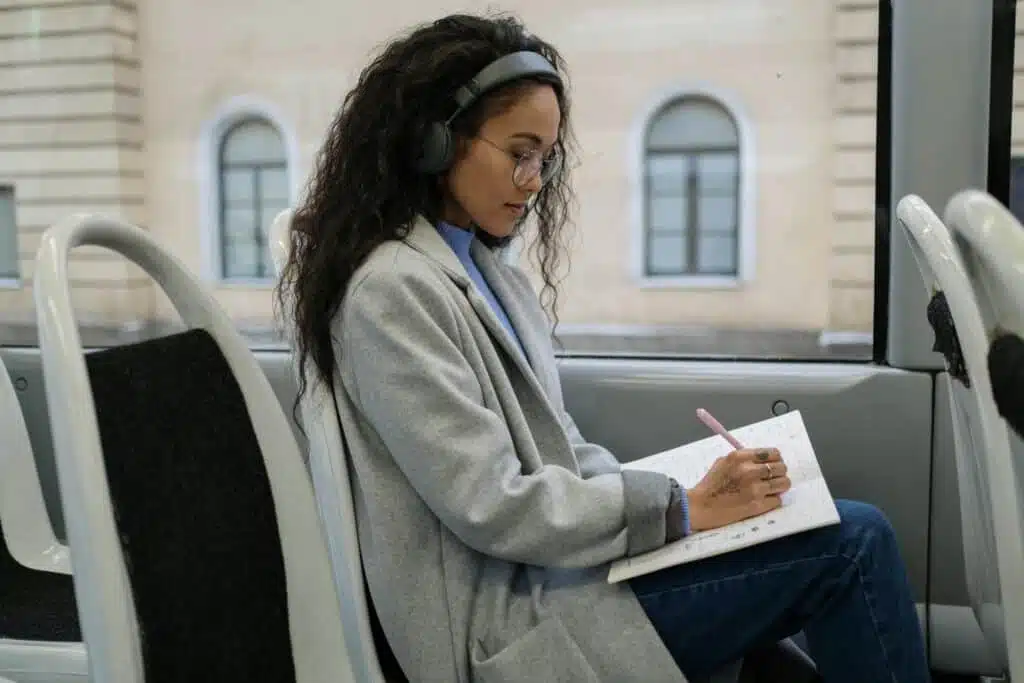 A woman with eyeglasses and headphones writing in a notebook while sitting on a bus.