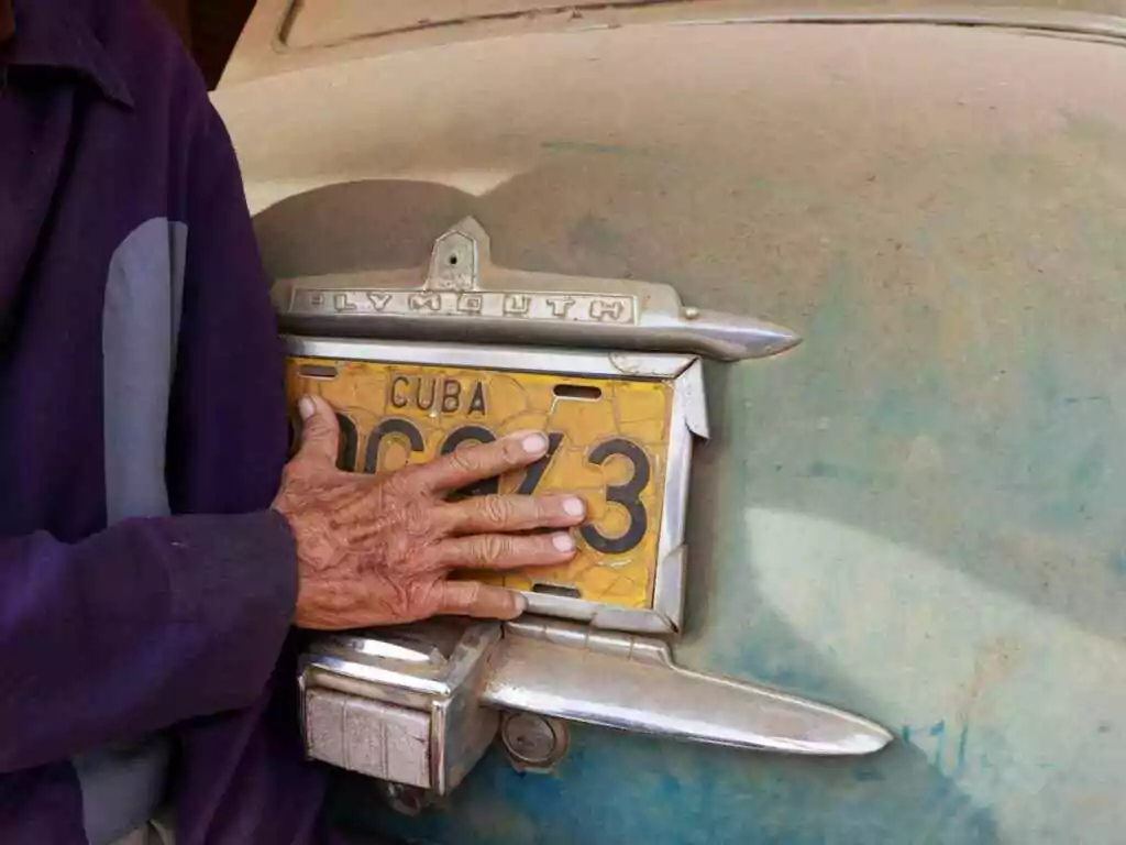 An elderly man's hand on a dirty vintage Cuban car's license plate.