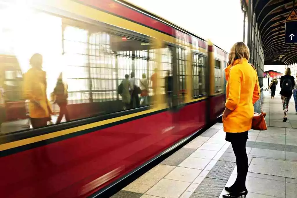 A woman in a yellow coat waits as a red train passes in a bustling station.