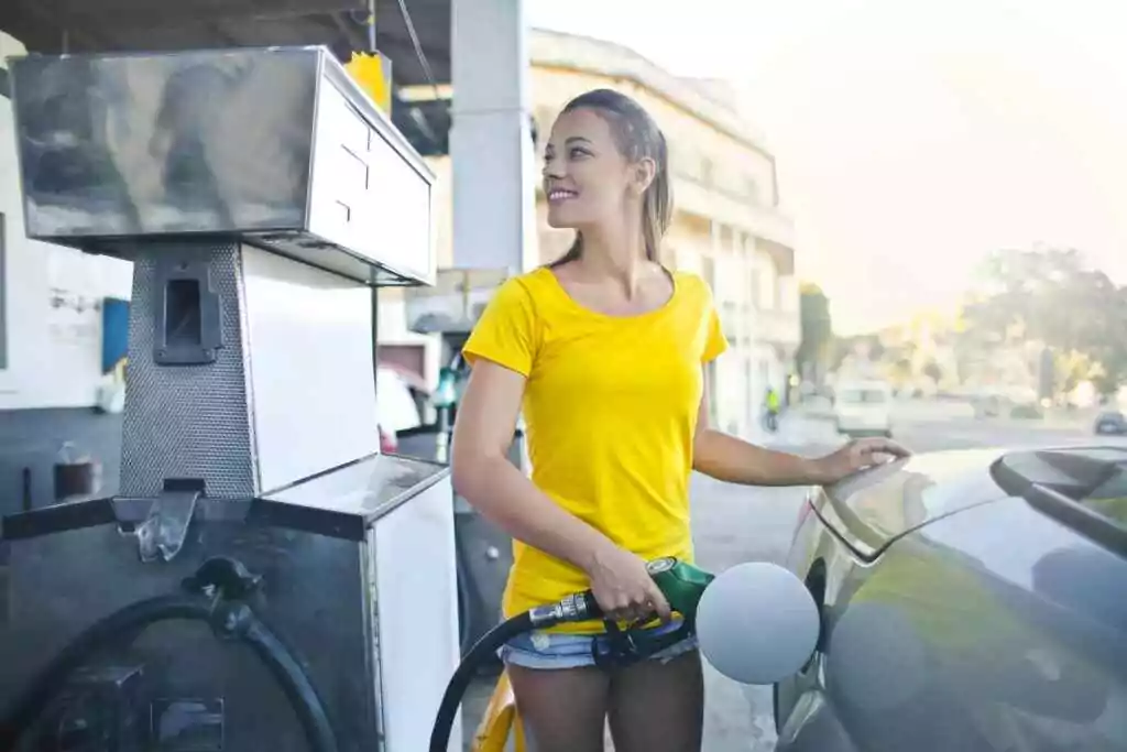 A smiling woman in a yellow shirt refuels her car at a gas station, enjoying a sunny day.