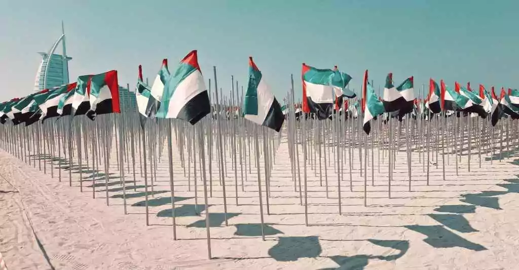 صفوف من أعلام دولة الإمارات العربية المتحدة على شاطئ مشمس في دبي، مع برج العرب في الخلفية.