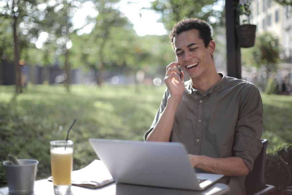 Man wearing casual clothes sitting at table in street cafe with laptop and speaking on cellphone