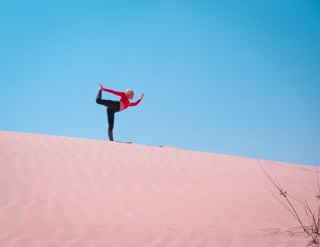 A woman practices yoga on sand dunes in Dubai, enjoying the clear blue sky and vast desert landscape.
