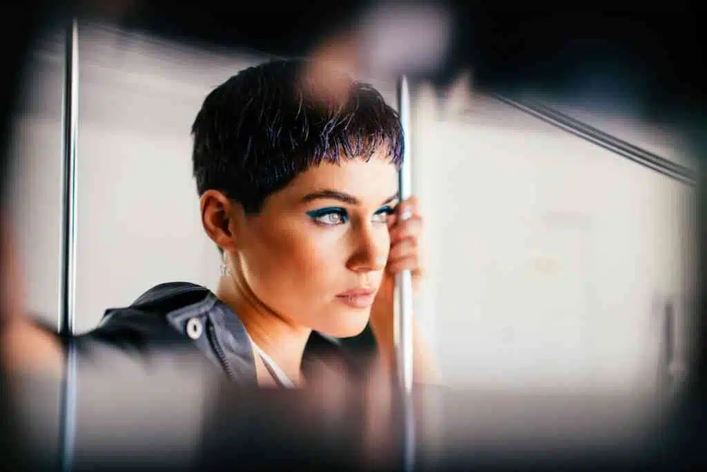 A young woman with short dark hair and bold makeup ponders while holding a railing in public transport.