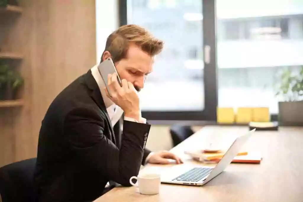 A businessman in a suit making a phone call while working on a laptop in a modern office setting.