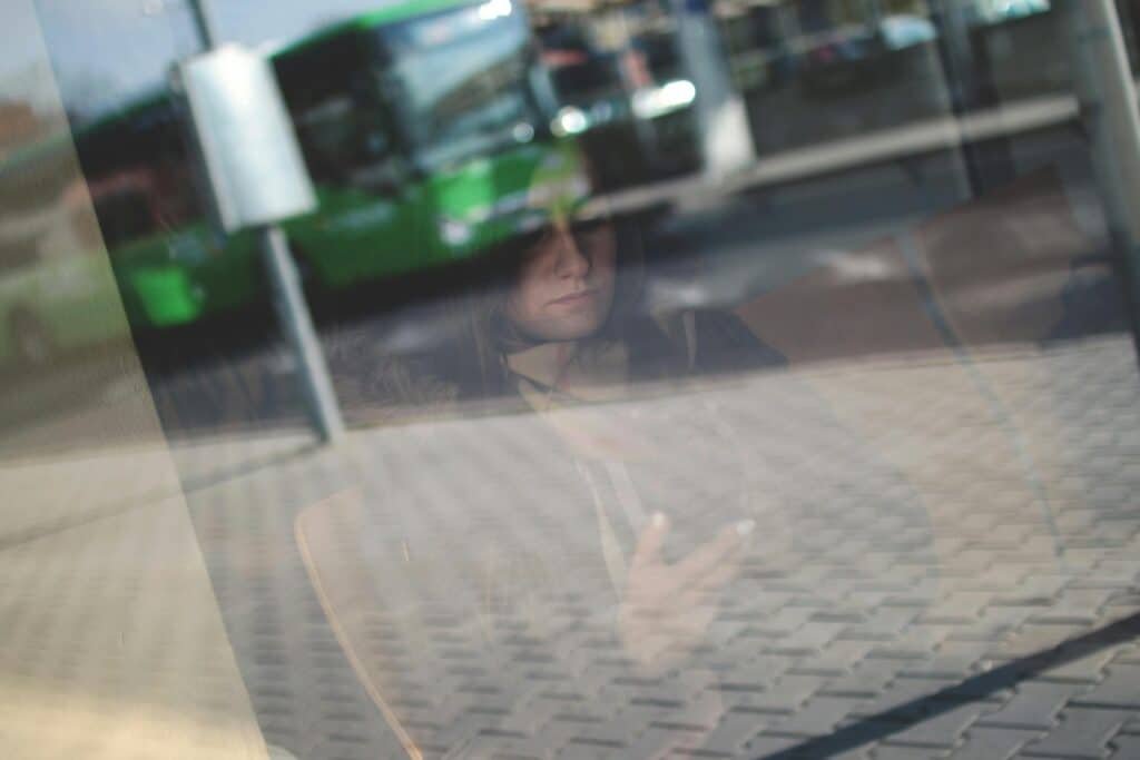 A woman is seen through glass using a smartphone in a waiting area, with a street reflection.