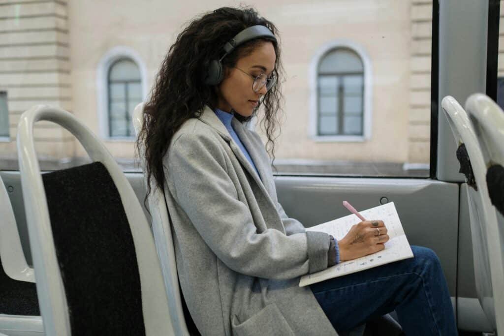 A woman with eyeglasses and headphones writing in a notebook while sitting on a bus.