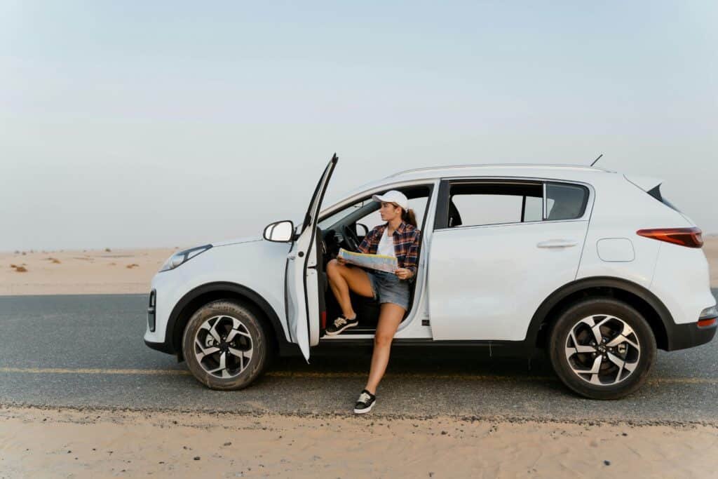 A woman sits by her SUV, exploring a map during a desert road trip under a clear sky.